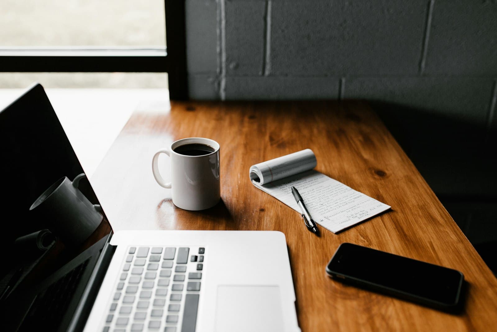 A laptop, coffee and a notebook on a wooden desk beside a bright window — a simple remote-work setup.