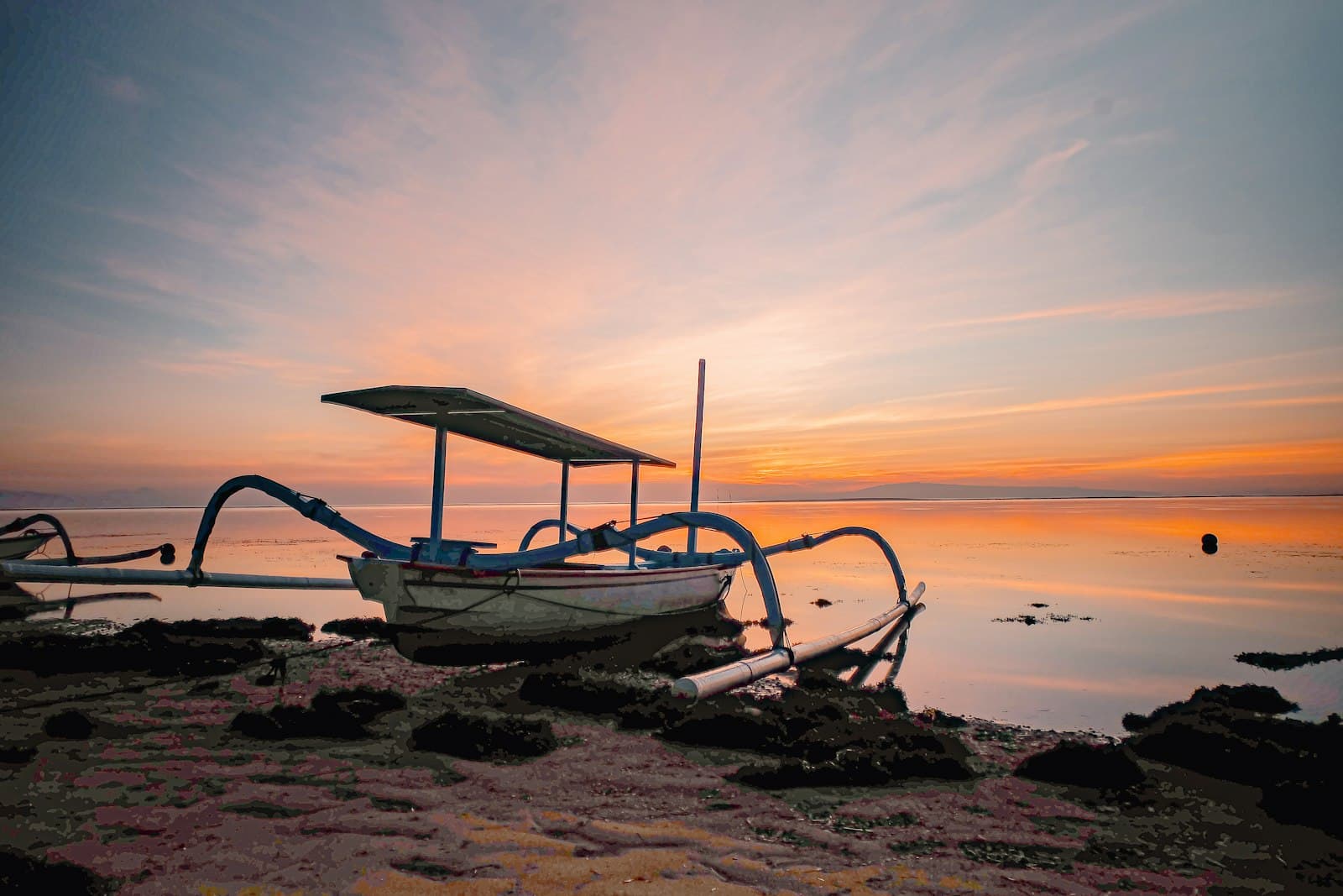 A traditional Balinese jukung outrigger boat on Sanur beach at sunrise, with calm water glowing orange.